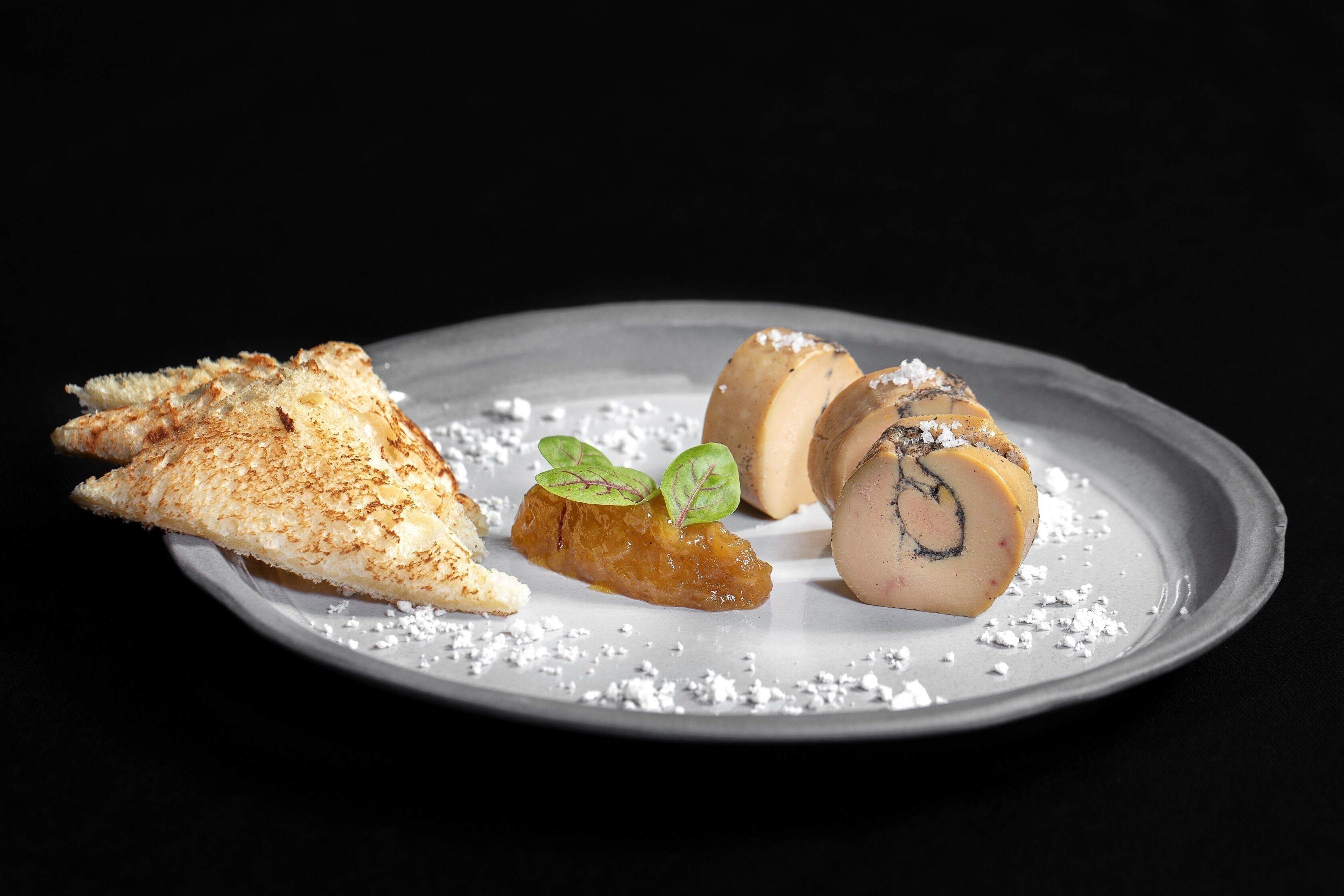 A plate with a piece of toasted bread, a rolled meat dish, and a small meat slice garnished with green leaves.