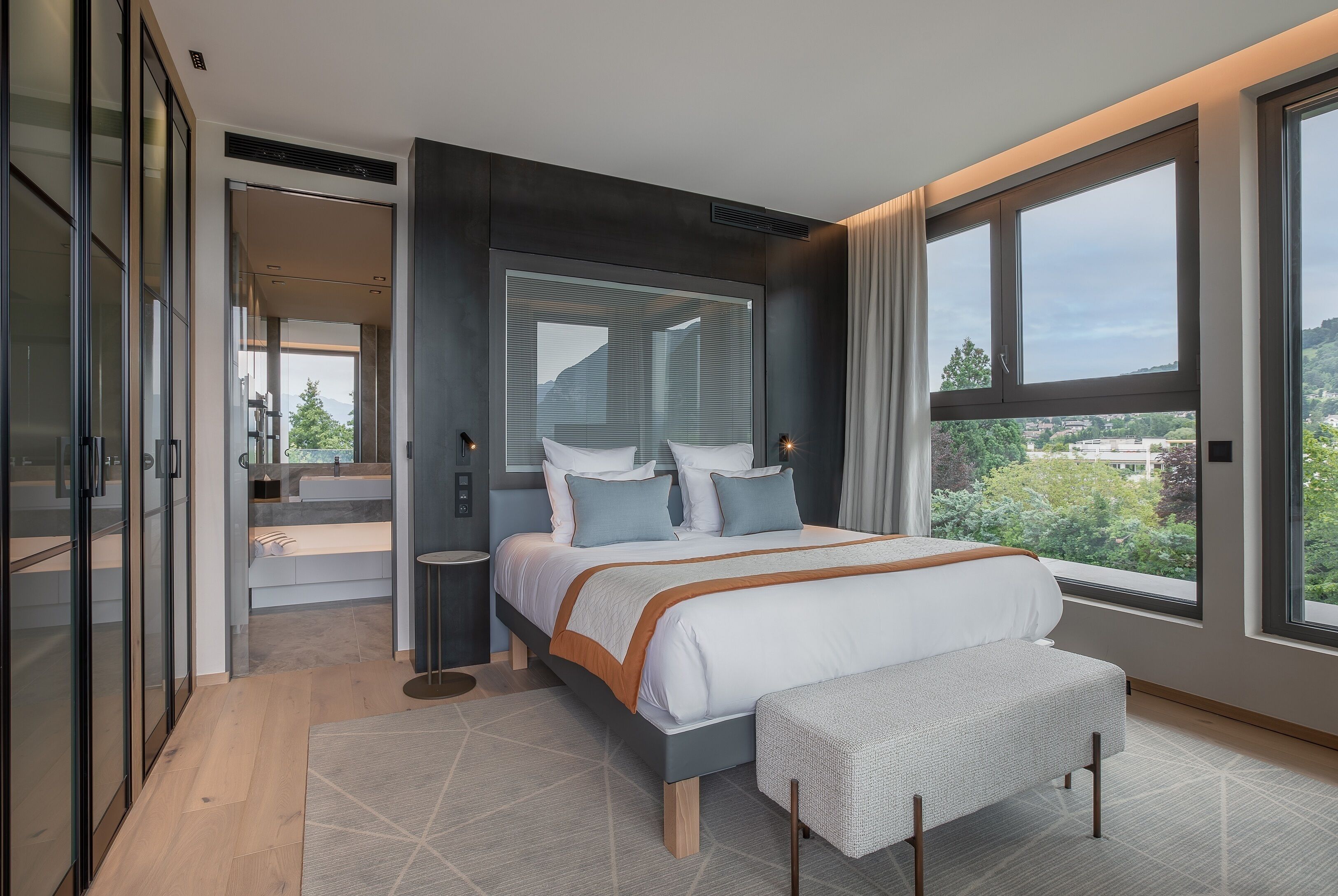 A modern bedroom with a large bed, a bench, and a view of the city.