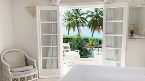 A bedroom with a bed, a chair, and a view of palm trees and the ocean through an open door. - Tradewinds, villa with sea views & pool, 1min walk to beach.  (Saint Peter)
