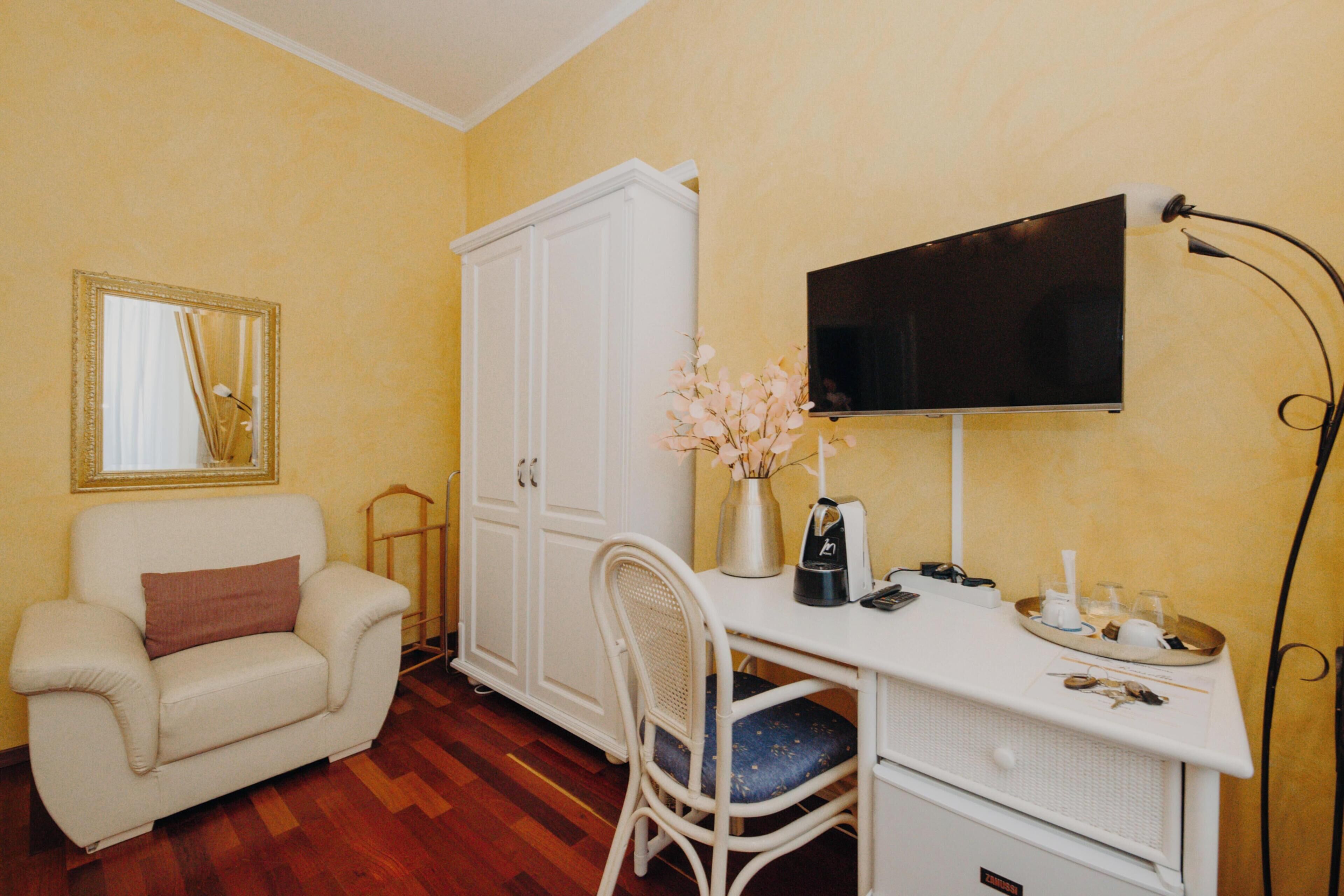 A room with a white desk, a chair, a television mounted on the wall, a wardrobe, and a mirror.