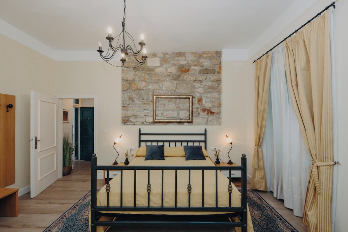 Une chambre avec un mur en pierre, un lit avec une tête de lit, un lustre et une grande fenêtre avec des rideaux.
