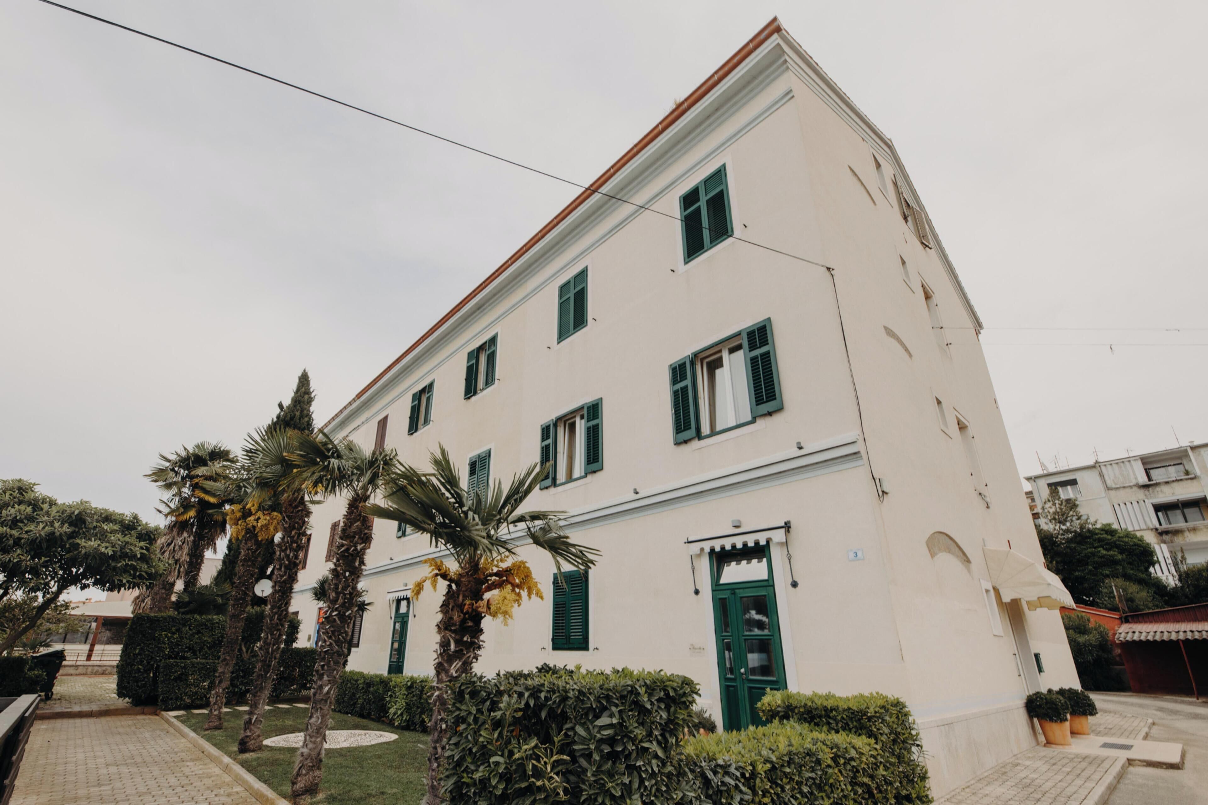 Un edificio blanco con contraventanas verdes, una entrada central y un patio empedrado con palmeras.