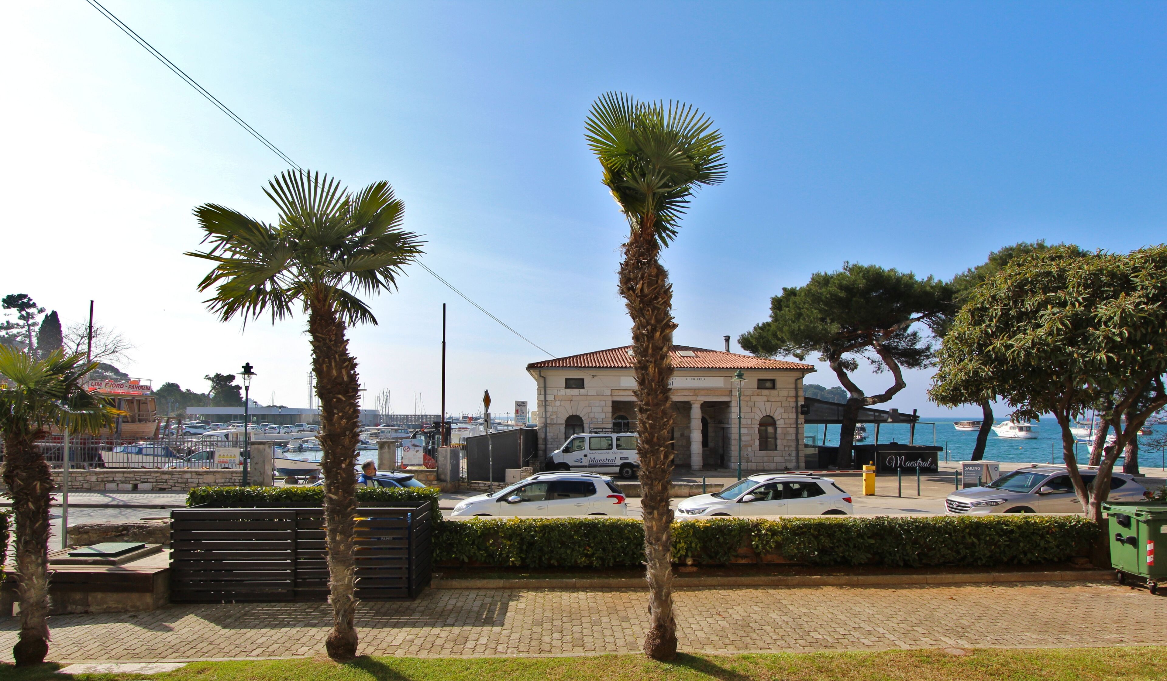 A coastal area with a stone building, parked cars, and palm trees.