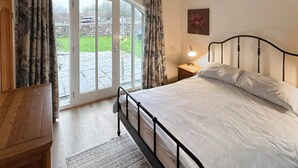 Ein Schlafzimmer mit einem Bett, einem Nachttisch, einer Lampe, einer Holzkommode und Blick durch eine Glastür nach draußen.