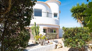 A two-story building with a white exterior and a curved roof. There is a paved pathway leading to the entrance, surrounded by gravel and small plants. - Exclusive villa with padel court [03] (Lagos)