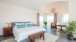 A bedroom with a large bed, a wooden bench, a nightstand with a lamp, a chair, a small table, and a potted plant. - Exclusive villa with padel court [03] (Lagos)