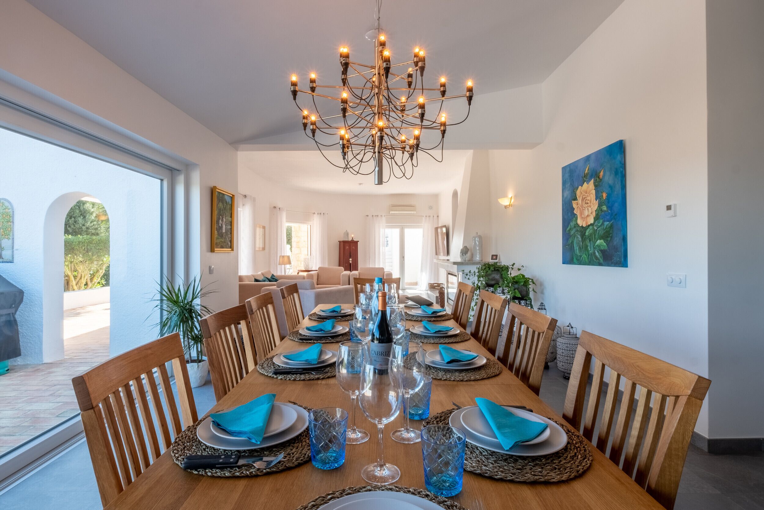 Un coin repas avec une longue table en bois dressée pour le repas, entourée de chaises en bois, d’un lustre et d’un tableau accroché au mur.