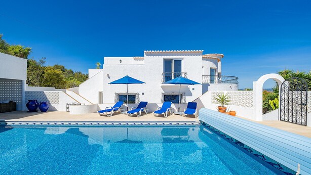 A pool area with blue lounge chairs and umbrellas, a white building with a balcony, and a clear blue sky. - Exclusive villa with padel court [03] (Lagos)