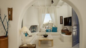 Un salón acogedor con un sofá blanco, una mesa de centro de madera, una lámpara y vistas a través de una ventana con cortinas blancas.