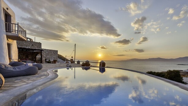Una piscina infinita con vista al atardecer sobre el mar, arquitectura de piedra y zona de estar al aire libre.