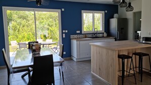 A modern kitchen with a large island, a dining area with a table and chairs, and a view of greenery through the windows.