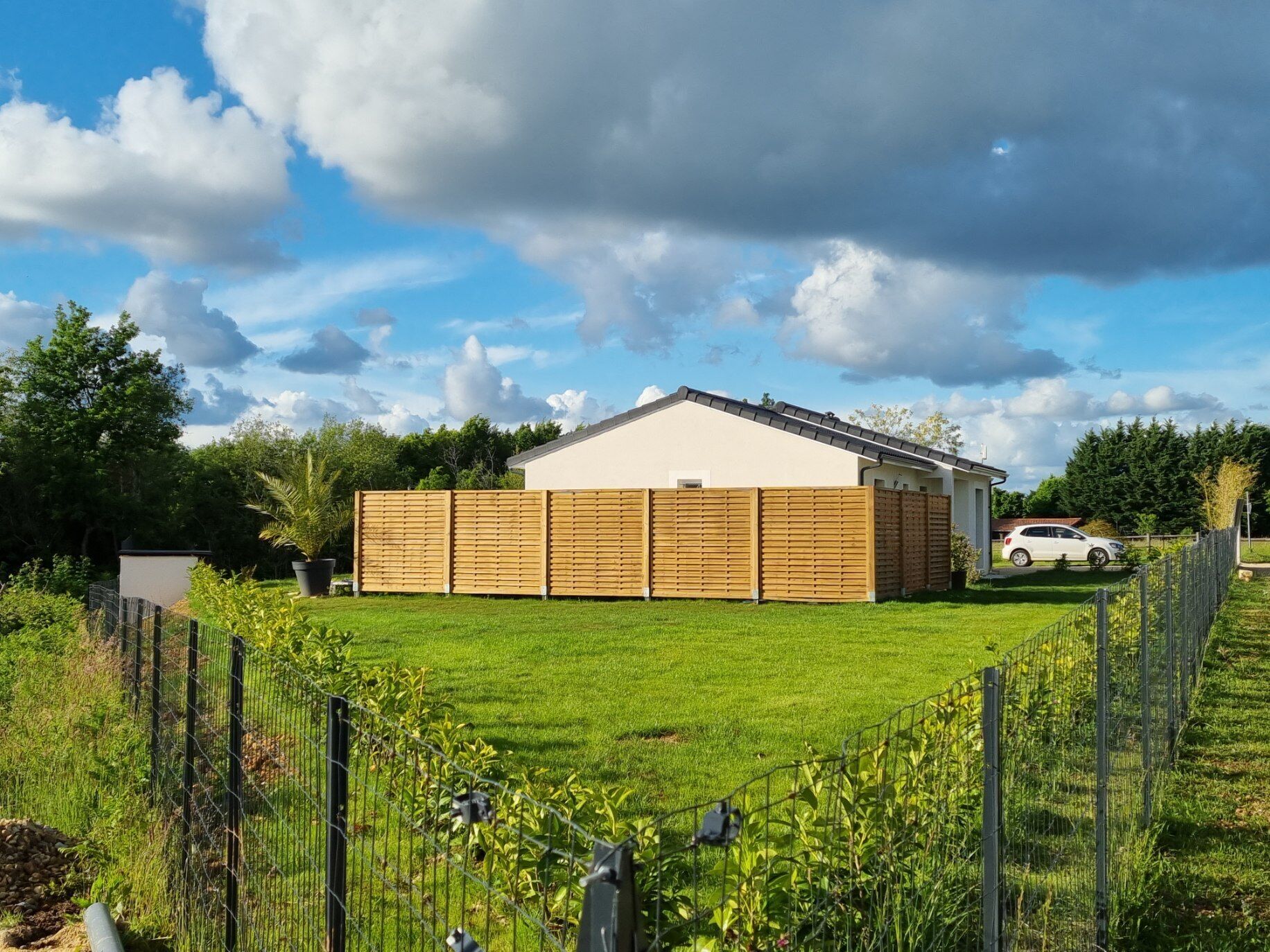 A residential property with a fenced backyard, a white car parked in the driveway, and a clear sky with clouds.