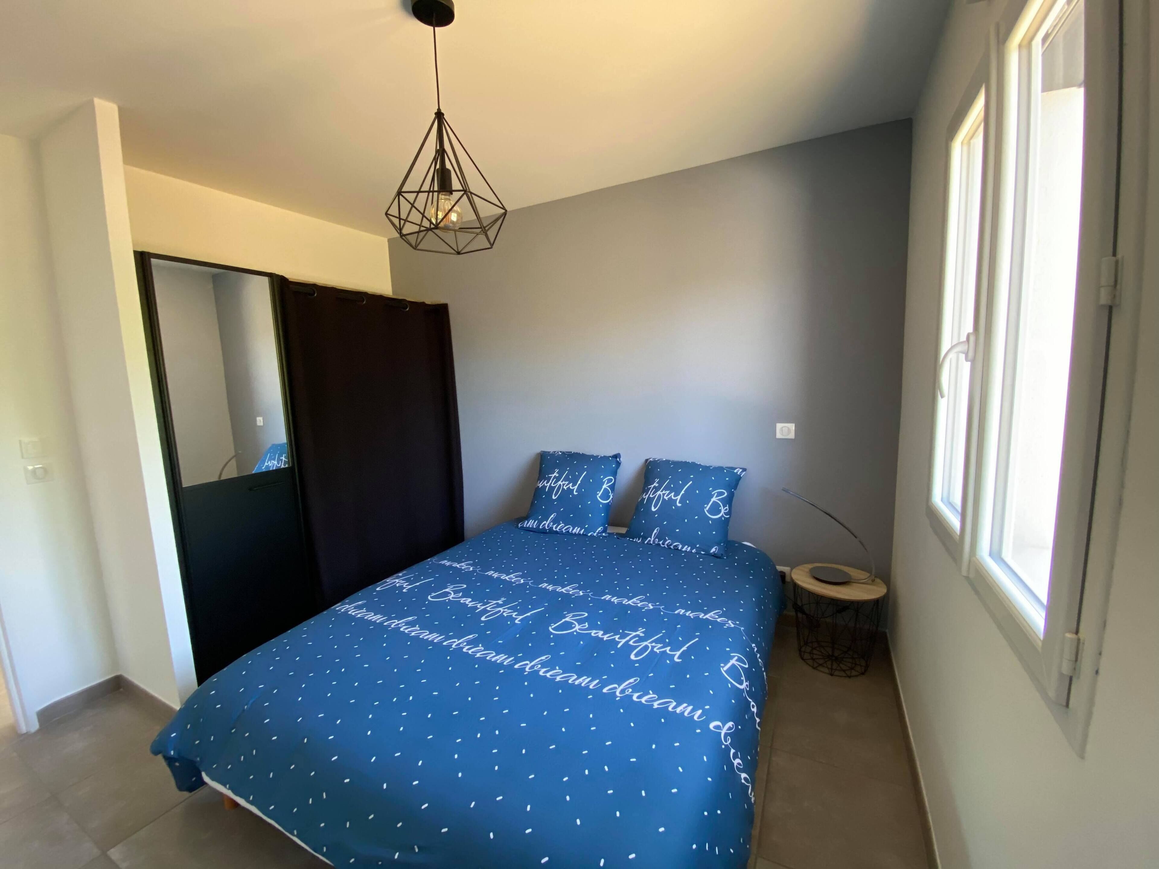 A bedroom with a bed, a mirror, a window, and a hanging light fixture.
