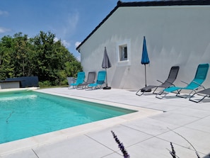 Una piscina con sillones de descanso y sombrillas, rodeada por un edificio blanco y árboles.