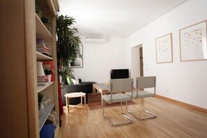 A modern living room with a sofa, a TV, a dining table, and a chair. There is a bookshelf with books and decorative items, and a large potted plant.