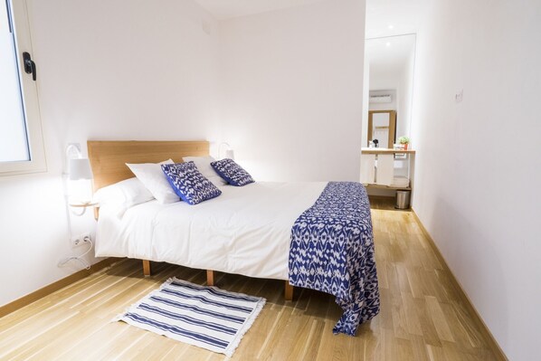 A bedroom with a bed, bedside lamps, a bedside table, a window, a mirror, and a wooden floor.