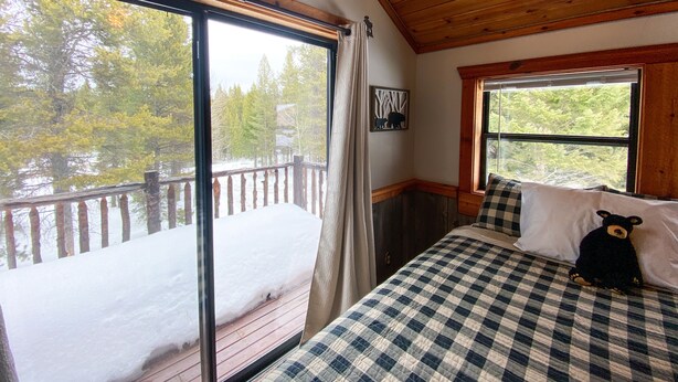 Una habitación acogedora con techo de madera, un ventanal con vistas a un porche nevado y árboles, una cama con colcha de cuadros y una almohada con motivos de oso.