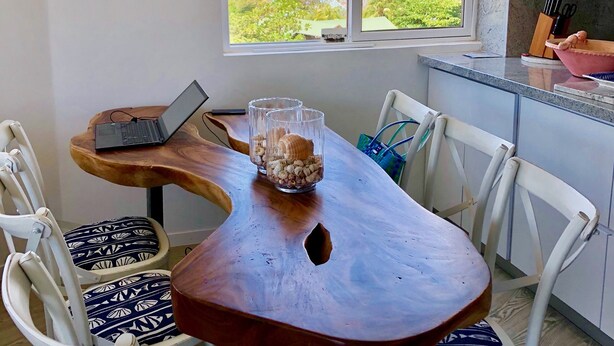 Zona pranzo con tavolo in legno, sedie bianche e un portatile. Una finestra offre vista sulla spiaggia e sugli edifici.