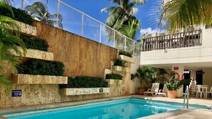 Una piscina con bordo piastrellato, circondata da un muro di legno e vegetazione. Sono presenti sedie a sdraio e un tavolino con sedie.