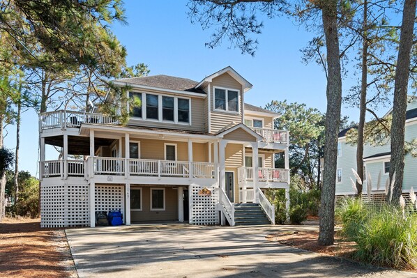 A two-story house with a balcony on the upper floor, a covered porch on the lower floor, and a driveway leading to the entrance. - Endless Sunsets in Corolla Light: Pool, Spa, Club Amenities & Golf Discounts! (Corolla)
