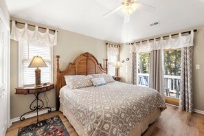 A bedroom with a large bed, a wooden headboard, two bedside lamps, a nightstand, a window with curtains, and a view of the outdoors. - Endless Sunsets in Corolla Light: Pool, Spa, Club Amenities & Golf Discounts! (Corolla)