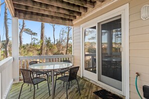 A covered outdoor deck with a round table and four chairs, a sliding glass door, and a view of trees and hills. - Endless Sunsets in Corolla Light: Pool, Spa, Club Amenities & Golf Discounts! (Corolla)