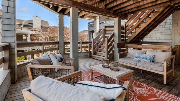 An outdoor wooden deck with a staircase, seating areas, and a view of surrounding buildings. - Carr'd Aweigh: Oceanside Condo, Shared Pool (Corolla)