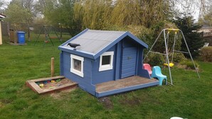 A small blue playhouse with a window and a door, a wooden play area with sand and toys, and a swing set in the background.