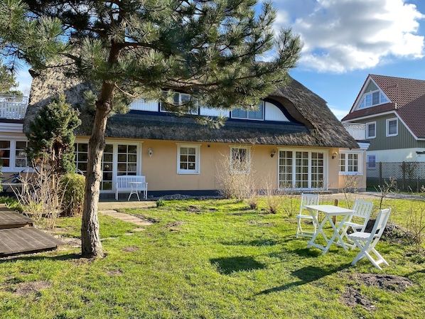 Une maison avec un toit de chaume, un petit jardin avec un banc et un arbre.