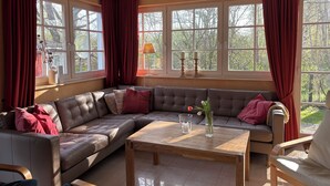A cozy living room with a brown leather sectional sofa, a wooden coffee table, and large windows with red curtains.