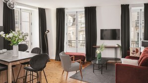 A modern living room with a dining area, large windows, a flat-screen TV, and a mix of furniture styles. - Appartements Crébillon - Maisons du Monde Hôtel & Suites (Nantes)