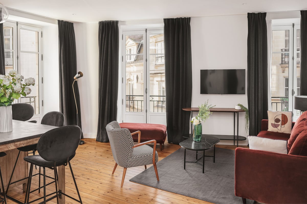 A modern living room with a dining area, large windows, a flat-screen TV, and a mix of furniture styles.