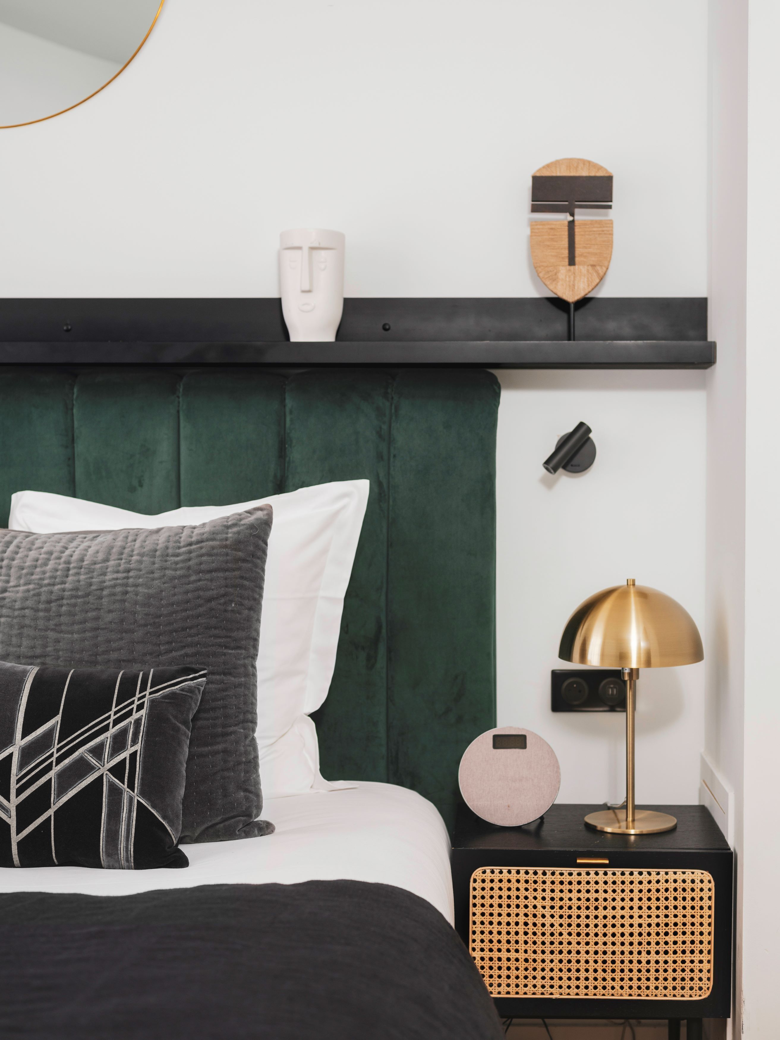 Un dormitorio ordenado con un cabecero tapizado en verde, una cama con ropa de cama blanca y negra, una mesita de noche con una lámpara y un estante montado en la pared con objetos decorativos.