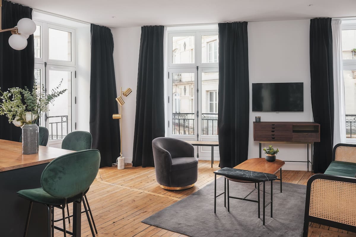 A modern living room with a wooden floor, a sofa, a chair, a small table, a TV, and a window with a view of buildings.