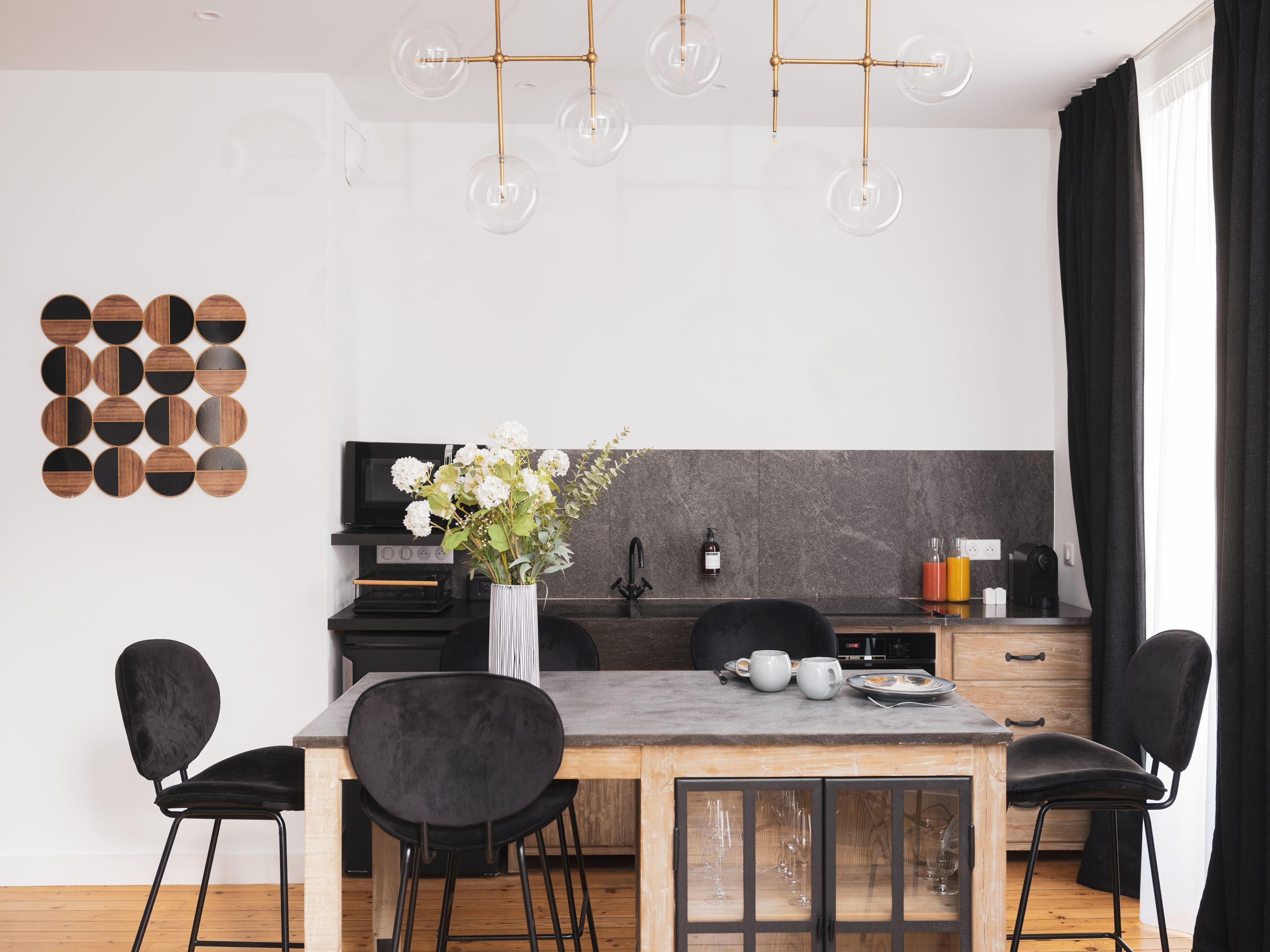 A modern kitchen with a wooden table, black chairs, a vase with flowers, and a hanging light fixture.