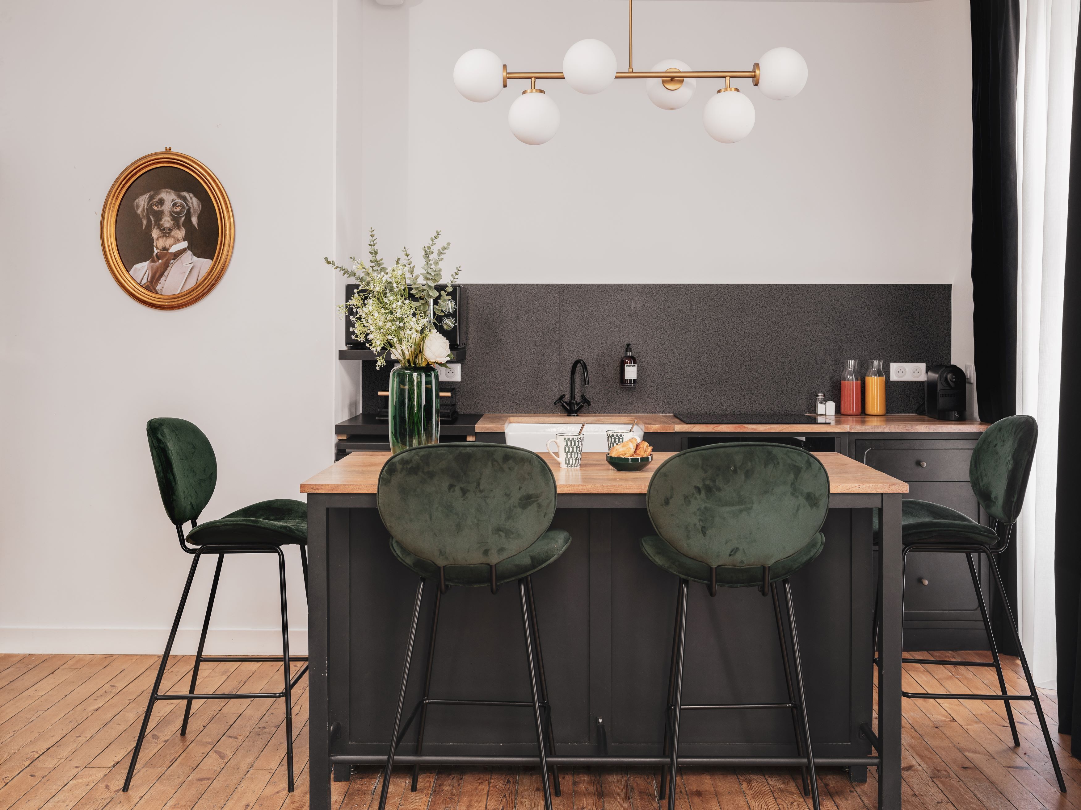 Una cocina moderna con una isla central, taburetes oscuros tipo bar, un jarrón verde con flores y un cuadro en la pared.