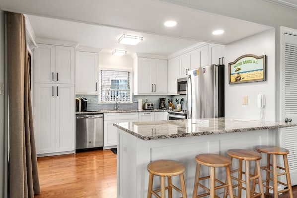 Una cocina con armarios blancos, encimera de granito, refrigerador de acero inoxidable, microondas y un lavabo. Hay tres taburetes de madera en la isla.