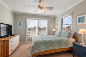 Une chambre à coucher avec un lit, un téléviseur posé sur une commode en bois, un ventilateur de plafond et deux fenêtres équipées de stores.