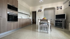 A modern kitchen with a built-in oven, a dining table with chairs, a fruit bowl, and a TV stand.