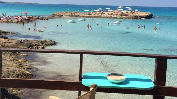 A beach with clear turquoise water, people swimming and sunbathing, and a rocky outcrop with umbrellas in the background.