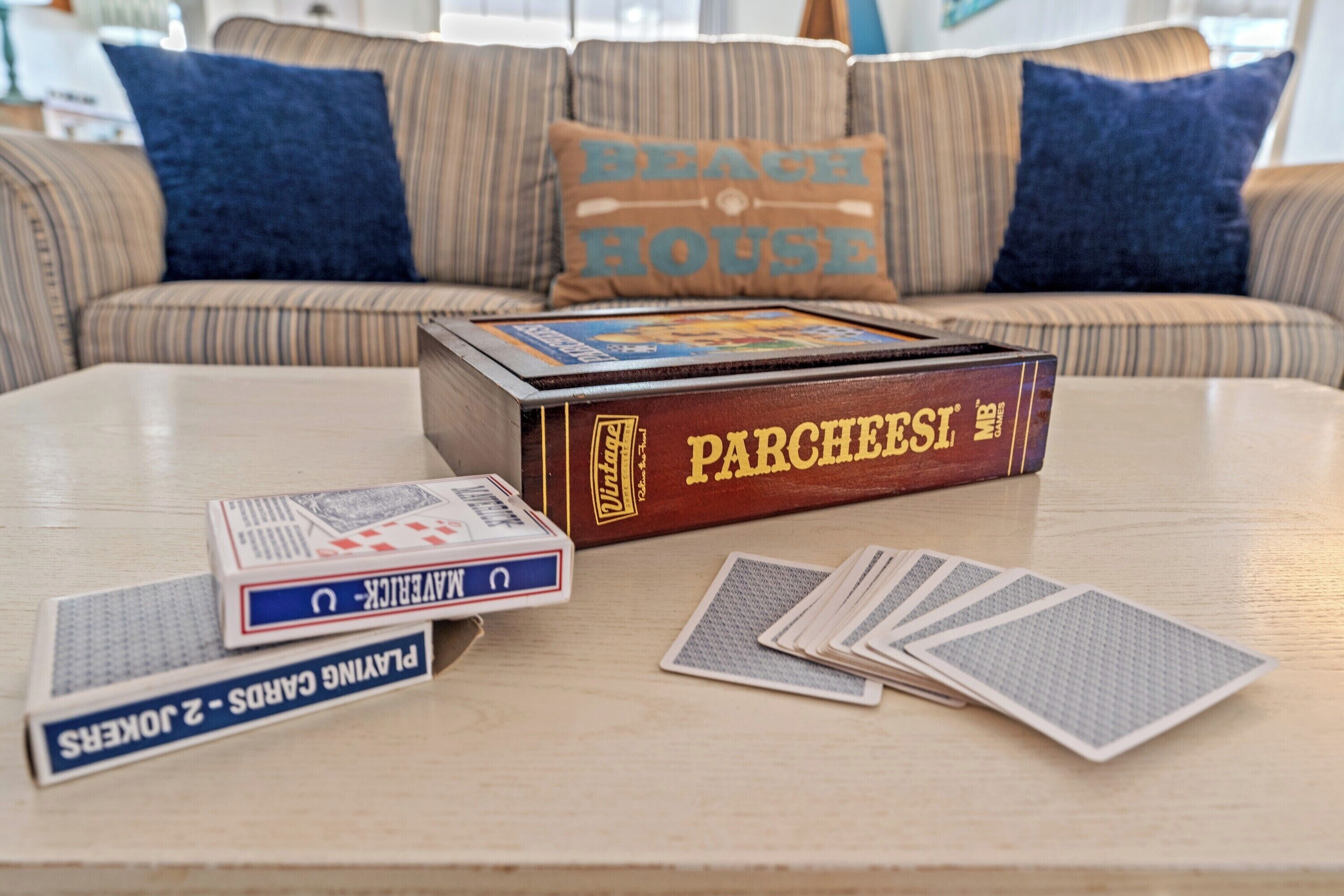 A game box labeled "Parcheesi" and playing cards on a table in a living room with striped sofas and a "Beach House" pillow.