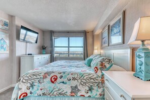 A bedroom with a bed, a dresser, a TV mounted on the wall, and a window with blinds.