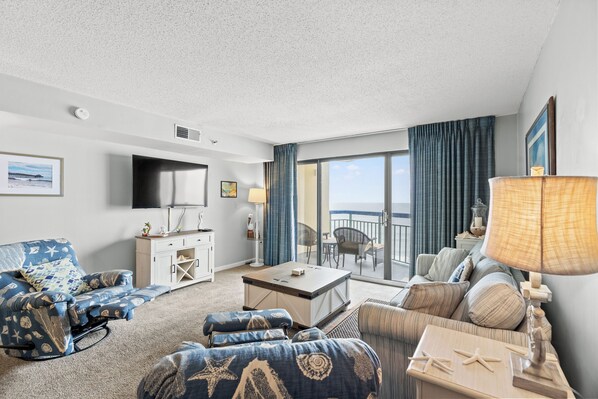 A spacious living room with a large flat-screen TV, a dining area, and a balcony with a view of the ocean.