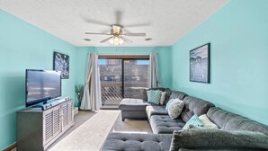 A living room with a large flat-screen TV, a grey sectional sofa, a ceiling fan, and a view of a building through the window.