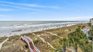 A beach with a wooden walkway leading to a building with a pool.