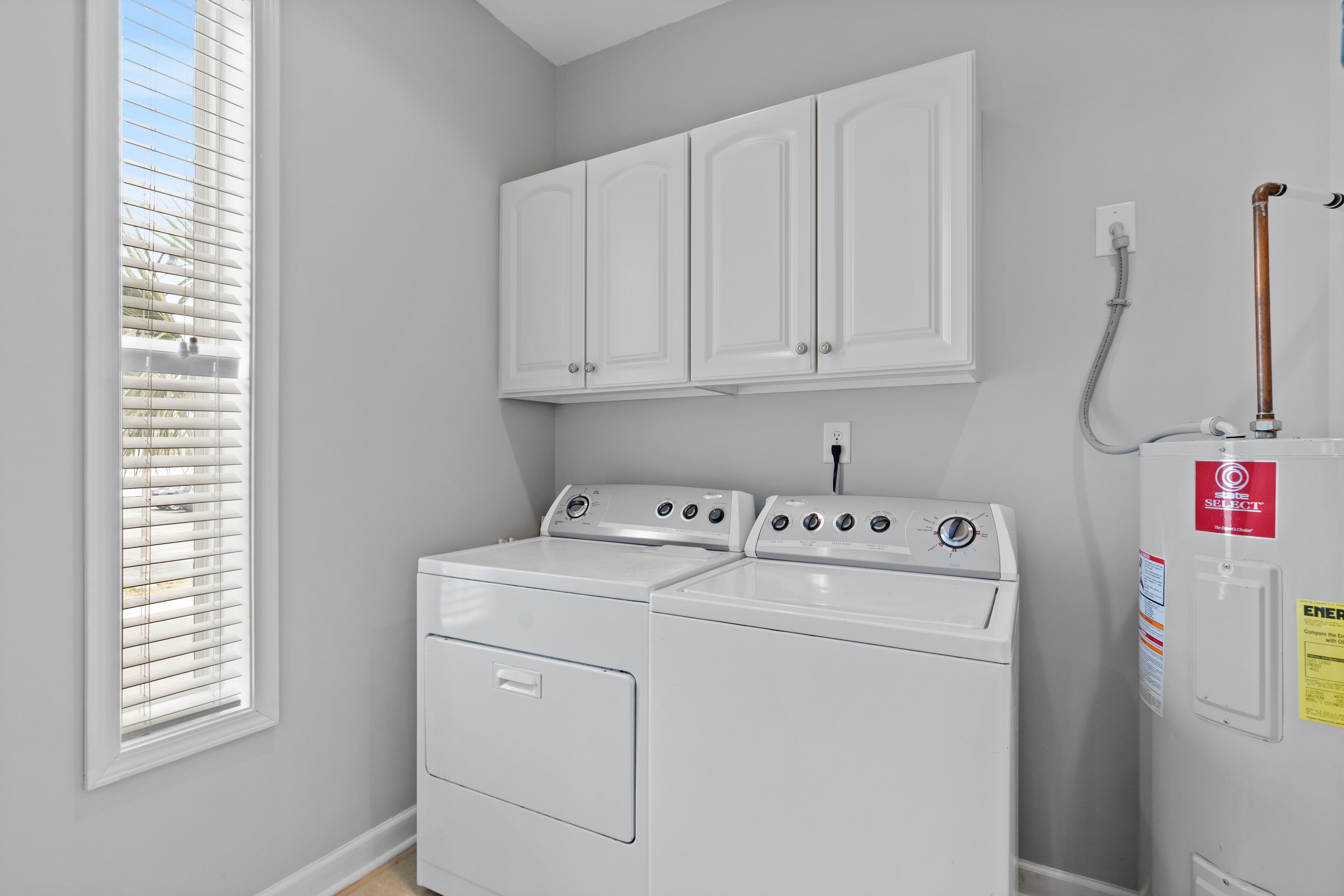 A laundry room with a front-loading washer and dryer, white cabinets above, and a window with blinds.