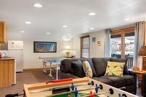 A living room with a foosball table, a sofa, a ping pong table, and a kitchen area.
