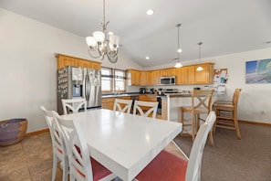 A modern kitchen with wooden cabinets, stainless steel appliances, a white dining table, and a chandelier.