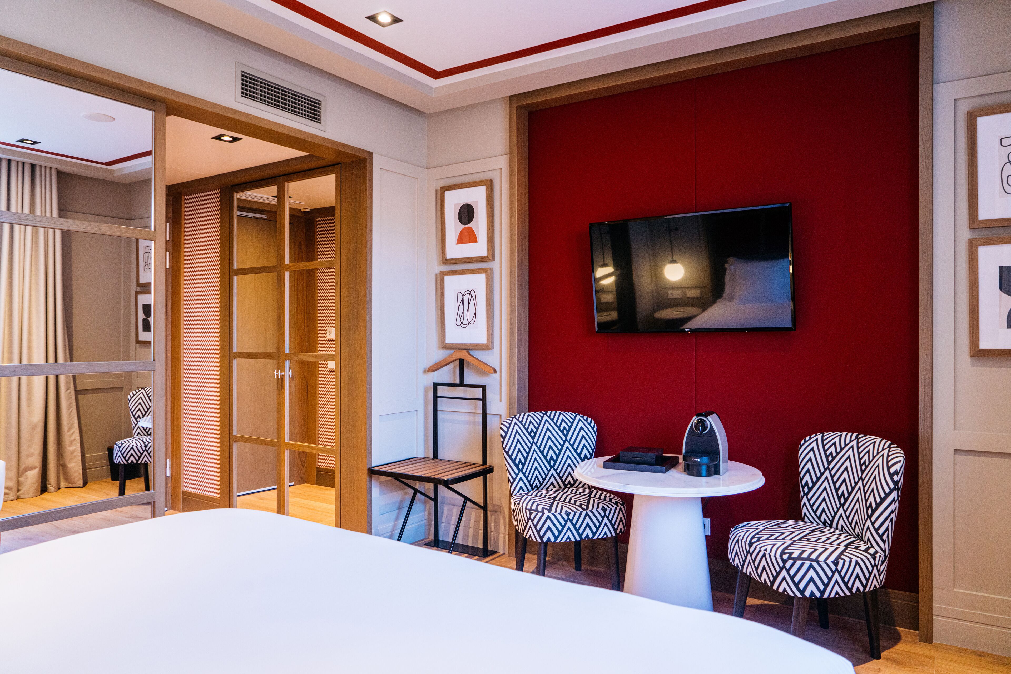 Ett hotellrum med en säng, en tv som är monterad på väggen, två mönstrade stolar, ett litet bord och en garderob i trä.