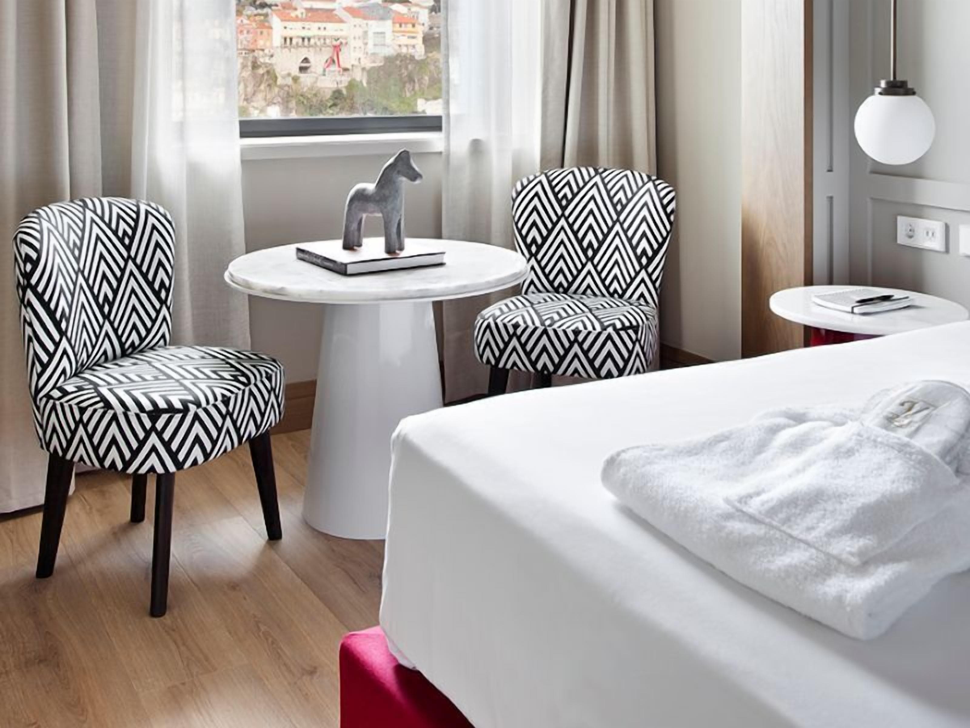 A hotel room with a bed, two patterned armchairs, a small round table with a sculpture, and a view of buildings through a window.